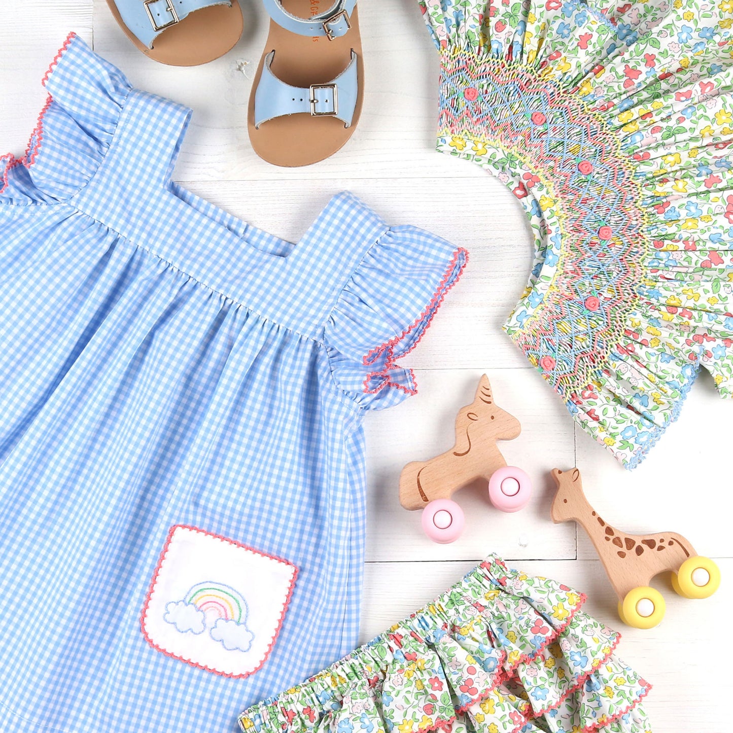 flatlay of Sunny Floral Smocked Bishop, light blue sandals, wooden toys and flower print dress and bloomers