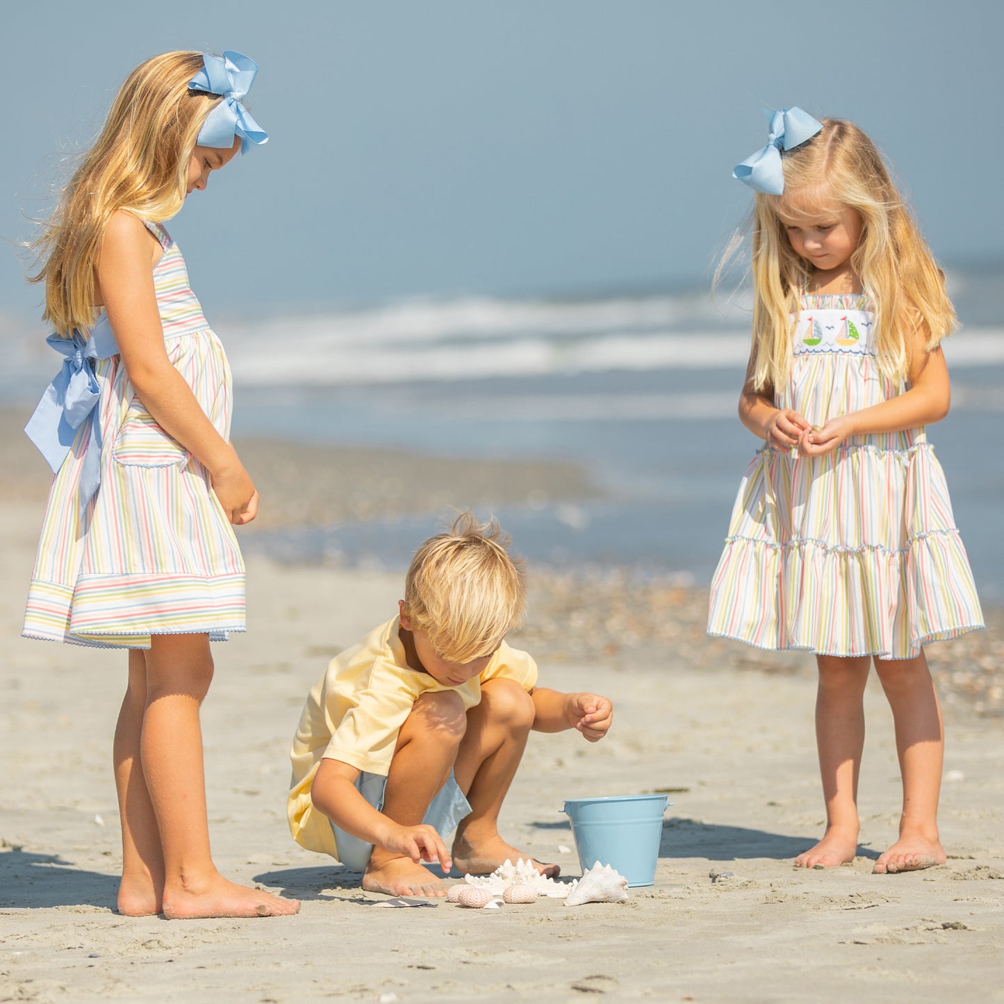 little girl wearing Yacht Club Stripe Smocked Dress picking up shells on the beach with a little boy and a little girl