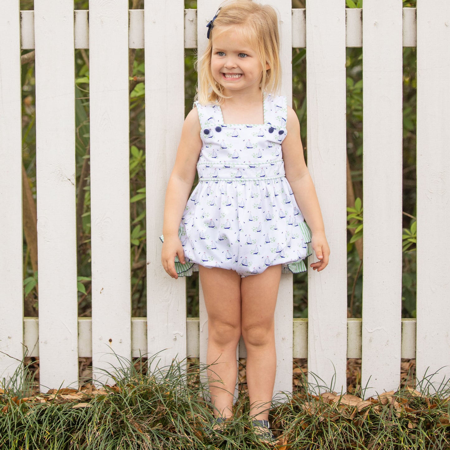 little girl wearing Beneteau Ruffle Bubble standing against a white fence