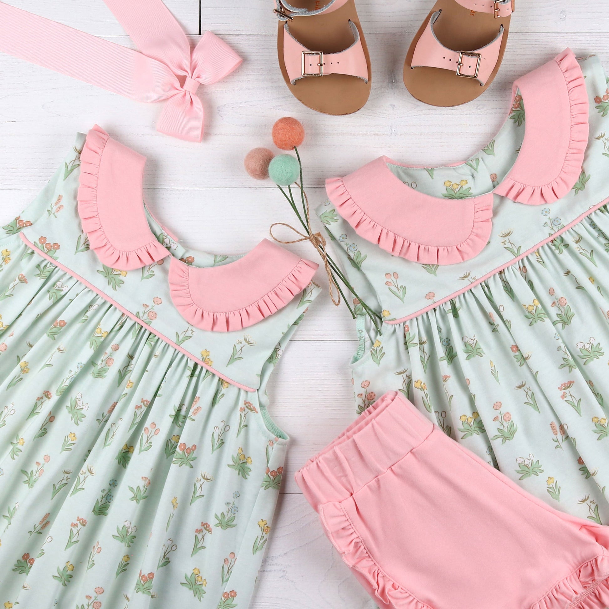 flatlay of a Girl's Secret Garden Dress and another dress, pink shorts, pink bow and pink sandals