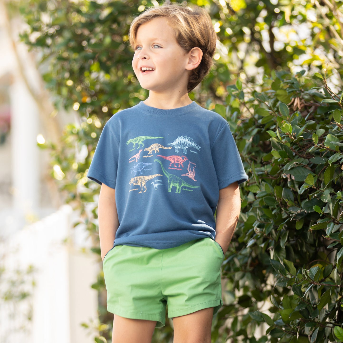 little boy smiling away from the camera standing next to a bush