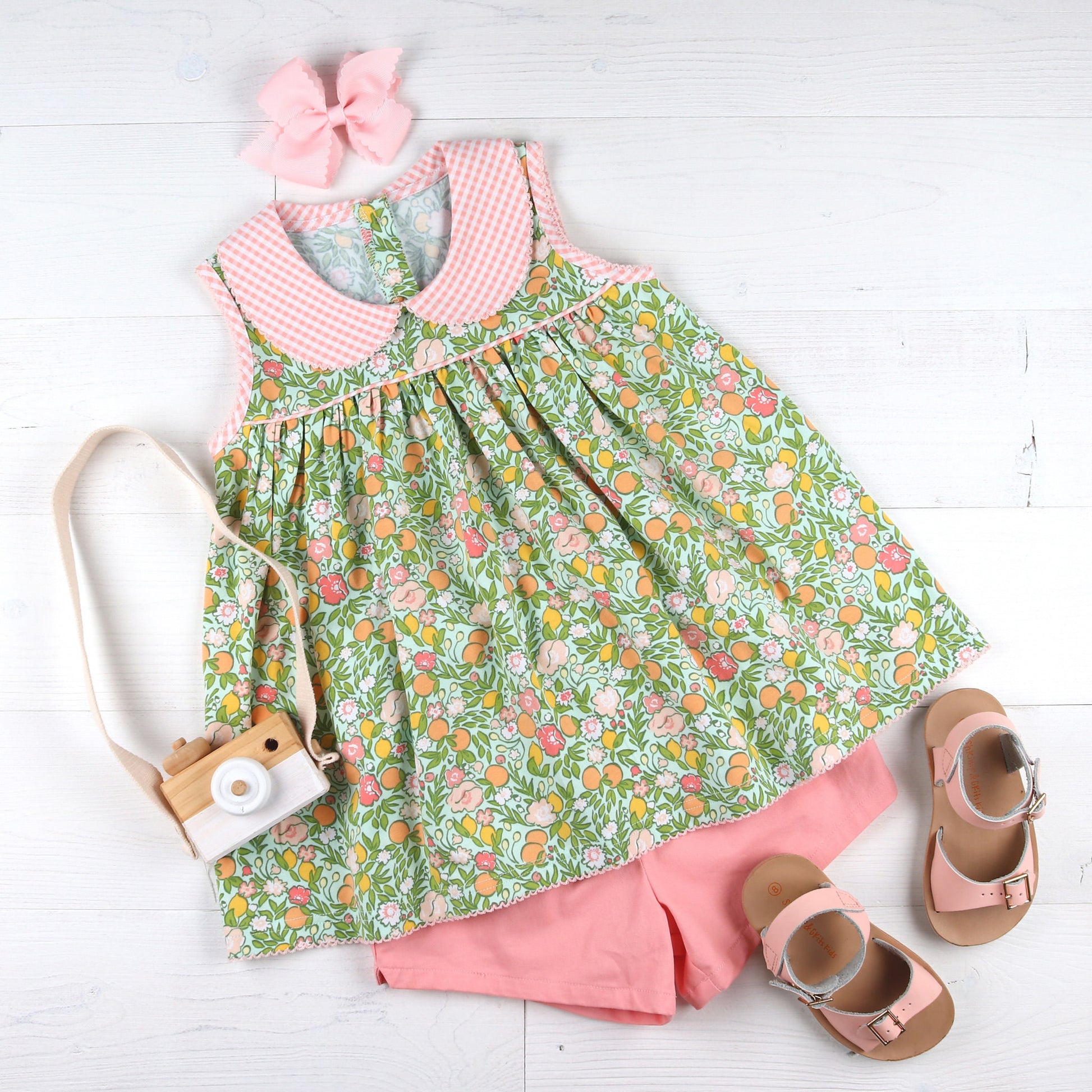 flatlay of flowerd print shirt, bow, sandlals and a toy camera