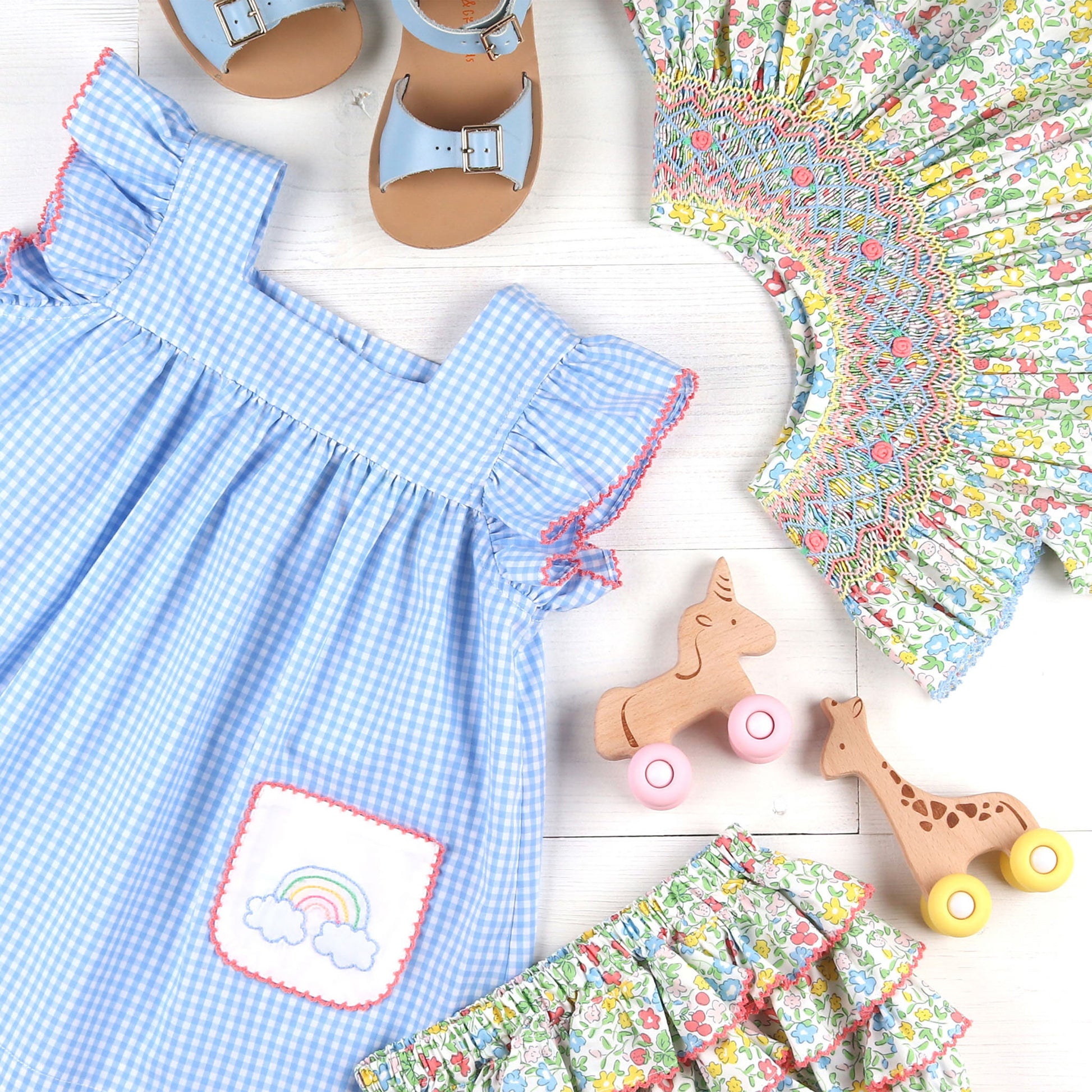 flatlay of Sunny Floral Smocked Bishop, light blue sandals, wooden toys and flower print dress and bloomers