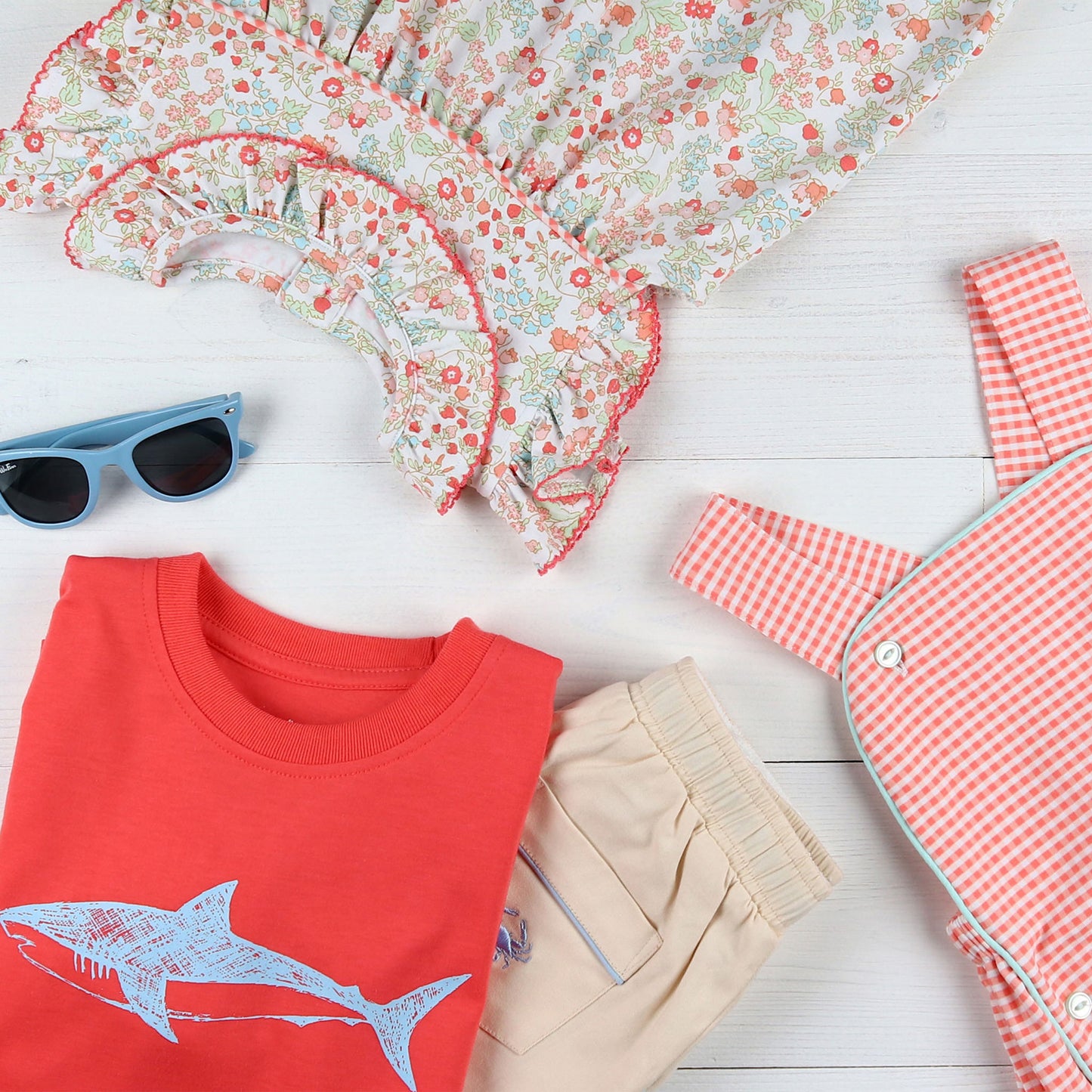 flatlay of a shark shirt, blue sunglasses, khaki shorts, checkered jumper and flower dress