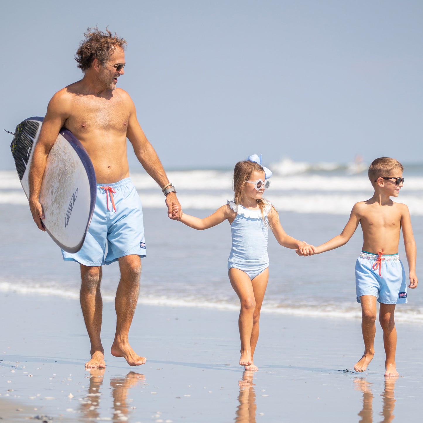 dad holding surfboard walking hand in hand with his kids wearing Skye Boardies