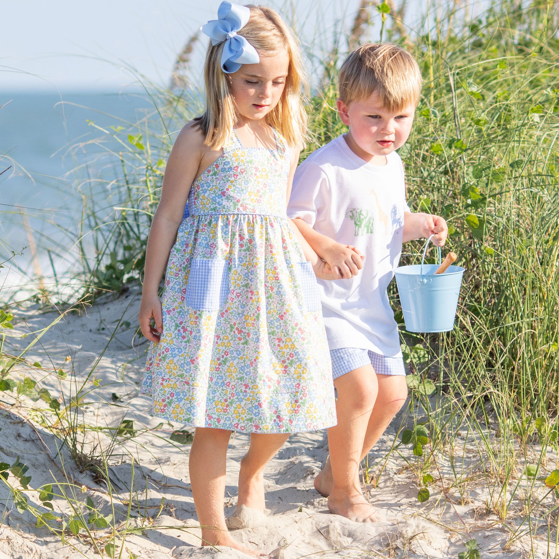 little girl in Sunny Floral Bow Back Dress and brother holding hands on the beach