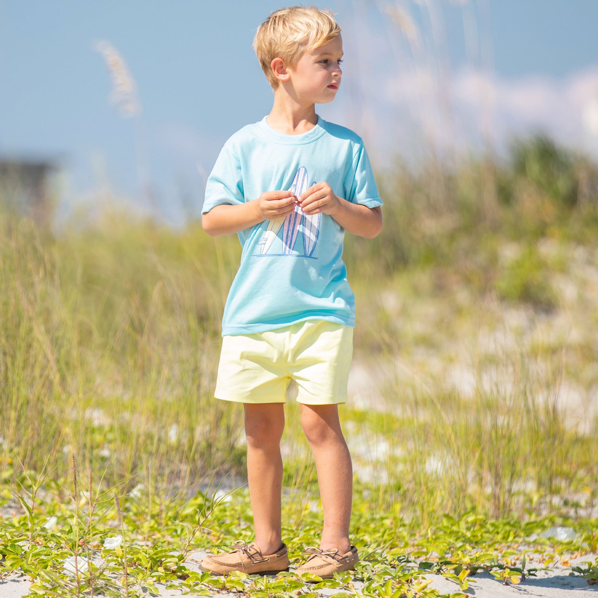 little boy standing on the beach wearing Surfs Up Graphic Tee and yellow shorts