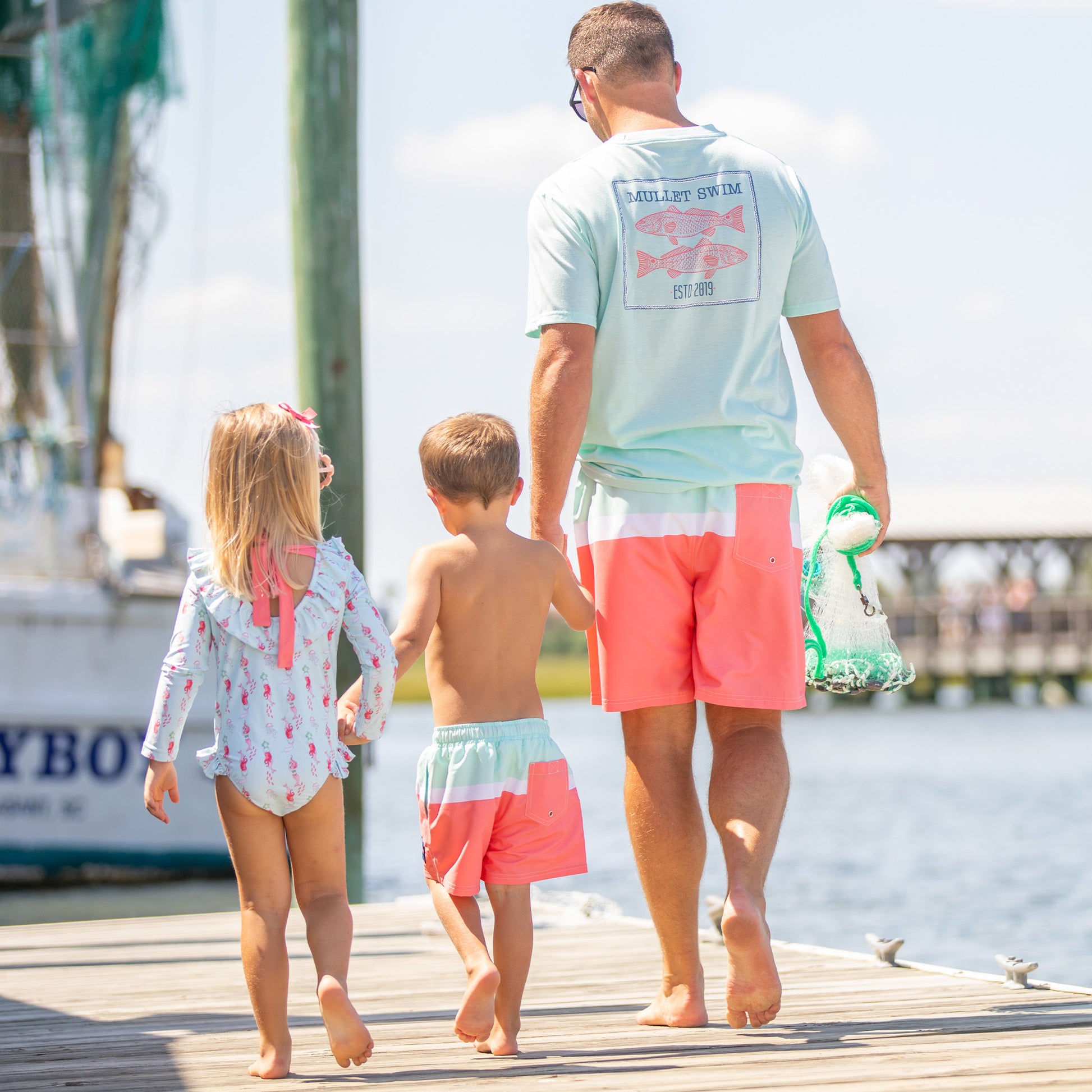 father wearing Red Drum Men's Graphic Tee holding hands with son and daughter on a boardwalk by the water