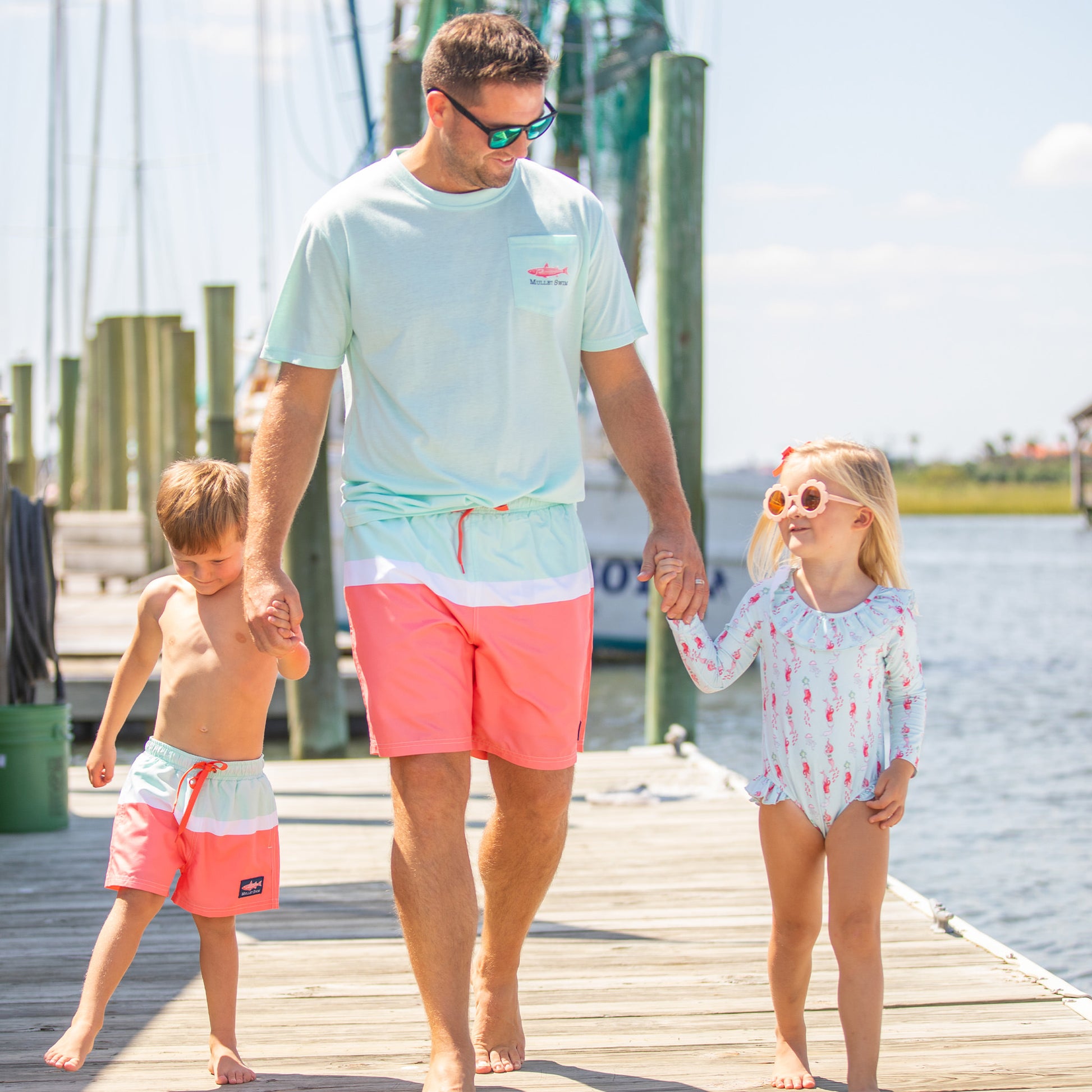 dad wearing a tshirt with a fish on the pocket holding hands with his son and daughter walking on the boardwalk on the creek